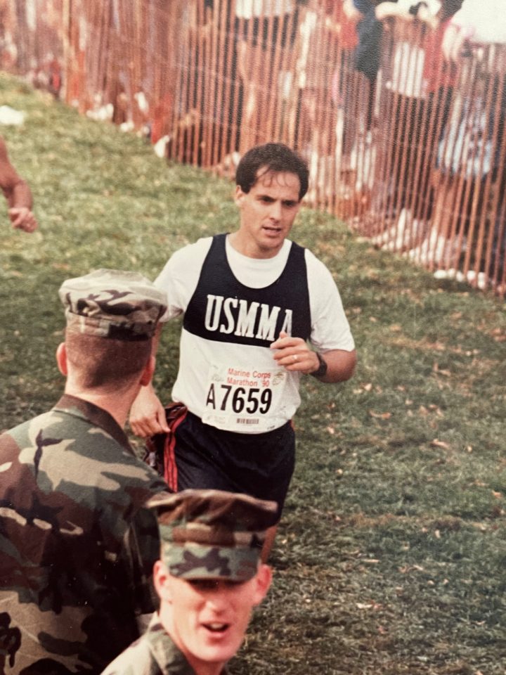 Man running a marathon as armed service members look on.